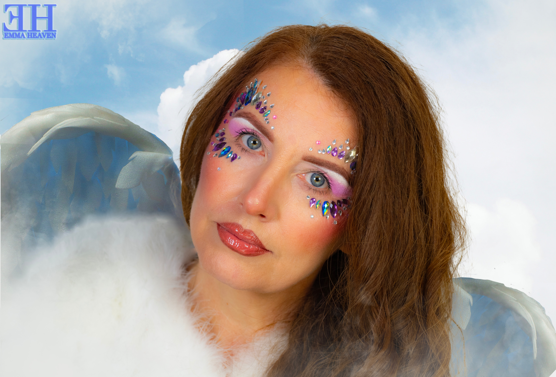 A woman with long brown hair and colourful makeup featuring sparkly gems around her eyes, wearing a fluffy white garment and angelic wings against a cloudy background.